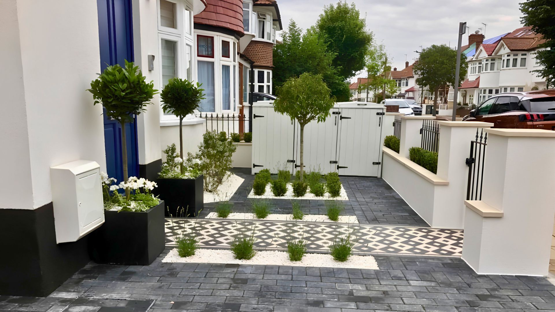 Charcoal block paving and white rendered front garden with clipped bay trees, lavender and a geometric black-and-white porcelain pathway leading to a bespoke timber bin store